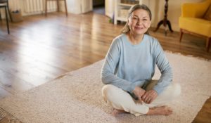 Indoor image of joyful happy middle aged woman wearing casual clothes sitting barefooted on white carpet at home